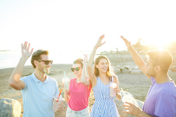 Group of happy teenage friends with drinks dancing on the beach on sunny day