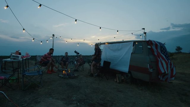 Parents with children holding skewers with marshmello and frying them in flame of bonfire having rest in campsite with van