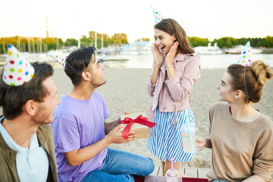 Surprised girl looking at her boyfriend with giftbox at birthday party on the beach