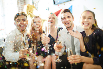 Young joyful friends with flutes of champagne having fun in confetti rain at birthday party