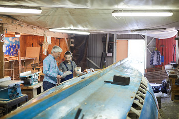 Young and senior workers discussing working instructions on paper while standing by unfinished vessel structure