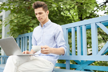 Fototapeta premium Young man with cup of coffee sitting on bench in summer cafe with laptop in front and searching for online ideas