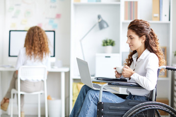 Young attractive woman in wheelchair having tea and reading online information in laptop