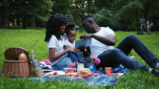 Portrait african american couple with daughter in city park young man woman and child doing picnic see photo album having fun outdoor slow motion smiling relationship summer kid nature together relax