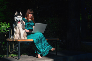 Beautiful young girl sitting on park bench with husky dog and laptop on her lap. Girl with long brown hair in long green dress. © Konstantin