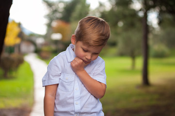 Portrait of a funny serious boy in a white shirt with a beautiful hairdo holding his chin with a hand against the background of blurry trees outside the city.