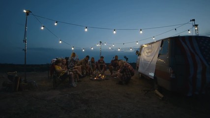 Group of cheerful friends with childrens relaxing in campsite and frying sausages on burning flame of bonfire gathering around and having fun