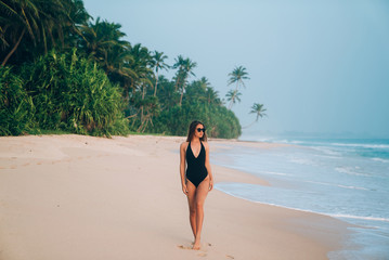 A beautiful young girl is resting on a tropical beach, against a background of greenery, palm trees, yellow soft sand and blue sea.Model in a stylish black swimsuit and sunglasses