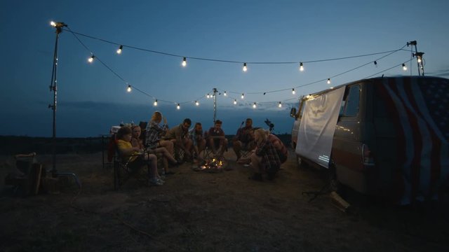 Group of cheerful friends with childrens relaxing in campsite and frying sausages on burning flame of bonfire gathering around and having fun