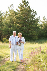Elderly couple in the white linen dress standing outdoors, in the hands a bouquet of flowers, and on the background of coniferous forest