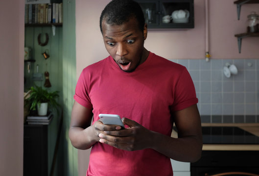 Closeup portrait of handsome African man standing in his kitchen holding mobile phone in both hands completely surprised with what he sees on screen experiencing extraordinary success or victory
