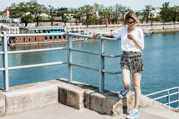 Stylish girl near the Vistula River in Krakow. Bridge across the river in Krakow. Young woman in town. Lady with a mobile phone. Travel to Poland. - obrazy, fototapety, plakaty