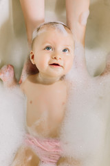 Happy laughing baby taking a bath playing with foam bubbles. Little child in a bathtub. Infant washing and bathing. Baby looking upwards in a plastic tub full of foam.