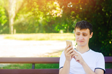 A young european guy sits on a bench in a city park and makes a funny face looking into the phone. The concept of extreme surprise and disgust, suddenness. Unexpected news, suddenness.