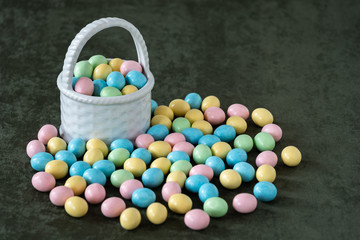 White ceramic basket on a dark green fabric background and pastel Easter candy eggs in yellow, pink, green, and blue
