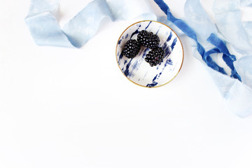 Feminine wedding or summer table composition. Porcelain plate with blackberry fruit and blue silk ribbons. Empty space. Flat lay, top view.