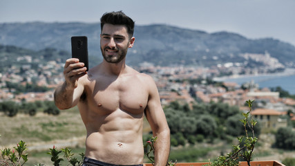 Young handsome man using smartphone to take selfie photo while standing shirtless on a balcony at...