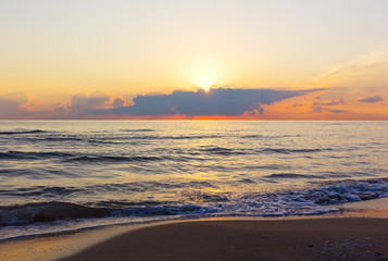 Sun appears above clouds during dawn over the Mediterranean Sea. Cloudy sunrise on an empty sandy beach.