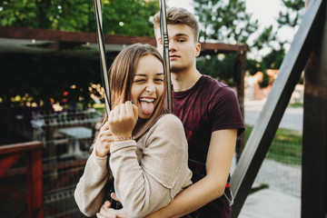 Obraz premium Amazing charming girl doing funny face showing her tongue with a closed eye while is embracing from back by her young man before ziplining on rope slide above the lake.