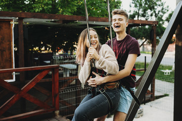 Handsome young man in summer clothes having fun laughing with his beautiful girlfriend while embracing her from back while hanging on a rope before ziplining.