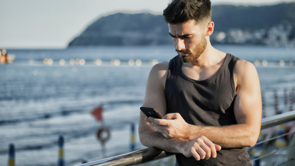 Athletic young man at the seaside using cell phone to type message while looking at the sea and...