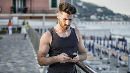 Athletic young man at the seaside using cell phone to type message while looking at the sea and beach