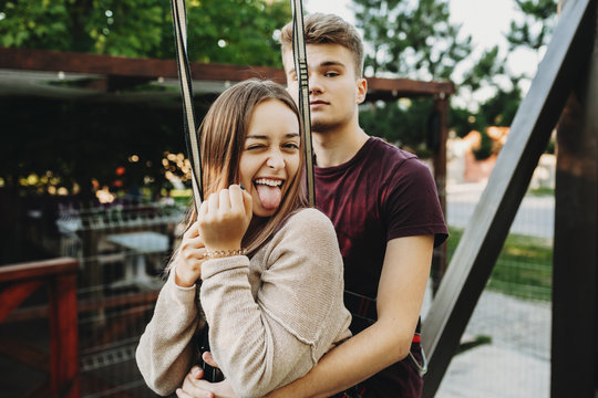 Amazing Charming Girl Doing Funny Face Showing Her Tongue With A Closed Eye While Is Embracing From Back By Her Young Man Before Ziplining On Rope Slide Above The Lake.