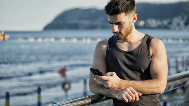 Athletic young man at the seaside using cell phone to type message while looking at the sea and beach
