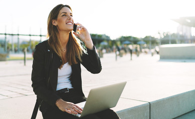Professional journalist female talking by a mobile phone with the editor while sitting outdoors with opened laptop on her knees. Freelancer making calls by a mobile phone while spending time outdoors.