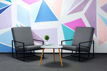 Two modern grey chair against a white wall with colored geometrical figure.Book laying on little table, green plant.Light of lamps.Copy space