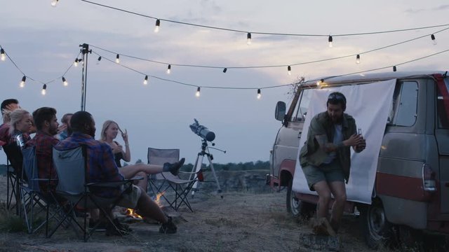 Group of diverse people chilling around bonfire in campsite and playing charades with woman acting and showing pantomime to others