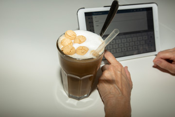 Person schreibt auf Tablet. Neben dem Tablet steht ein Glas mit Eiskaffee. Oben ist aufgeschäumte Milch und einige Amaretti. Außerdem ist ein durchsichtiger Strohhlam und ein langer Löffel im Glas.