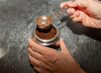 A person is putting coffee powder with a special spoon into a silvern coffee machine. 