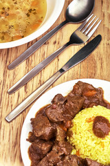 Plate with meat and liver under sauce with rice and plate with soup with fork, knife and spoon on wooden table