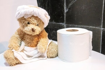 Bear wears towel on head and body with tissue paper is in toilet.