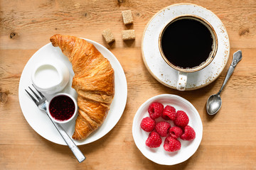 Croissant, cup of black coffee, fresh raspberries, cream and jam on wooden table. Continental breakfast, coffee break or breakfast concept. Table top view