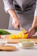 Pieces of pepper on a wooden board, female hands cut vegetable for salad on a white kitchen table
