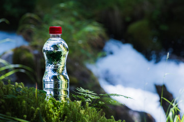 Transparent plastic A bottle of clean water with a red lid stands in the grass and moss on the background of a rugged mountain river. The concept of pure natural drinking water