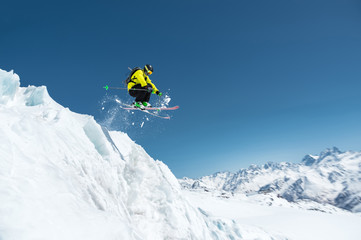 A skier in full sports equipment jumps into the precipice from the top of the glacier against the background of the blue sky and the Caucasian snow-capped mountains. Elbrus region. Russia