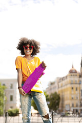 New purchase. Joyful curly woman showing her purple mini skateboard and smiling while posing for the camera in the downtown