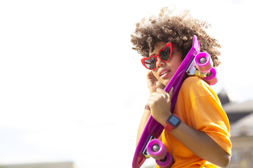 Urban fashion. Beautiful curly girl in heart-shaped sunglasses and a yellow t-shirt carrying a purple mini skateboard while looking down