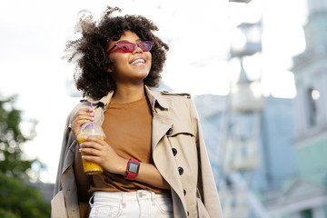 Bright day. Pretty fashionably-dressed young woman in a trench coat smiling happily and holding a glass of juice while walking down the streets