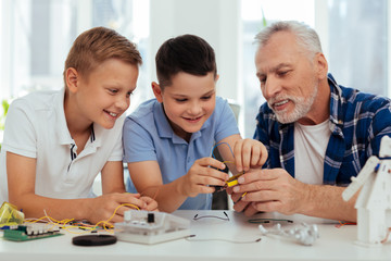 Pleasant hobby. Joyful smart children sitting together with their grandfather while having fun