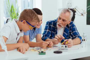 New skills. Nice smart man looking at his grandchildren while teaching them how to build robots