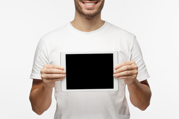 Studio picture of positive man isolated on grey background standing in casual clothes holding tablet and showing it blank screen with happy smile as if advising product or service