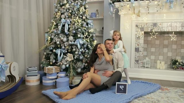 The little girl hugs and kisses her parents, dad and pregnant mom near the Christmas tree in the Christmas interior.