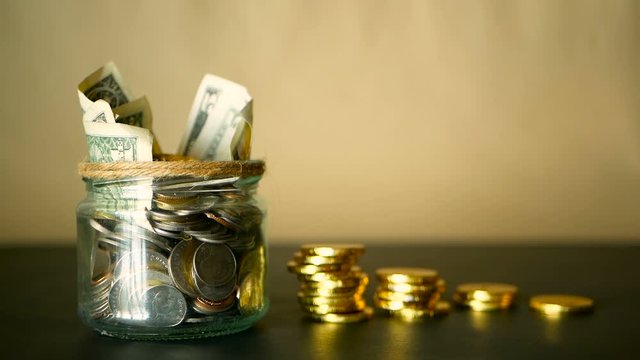 Saving money coin in jar. Symbol of investing, keeping money concept. Collecting cash banknotes in glass tin as moneybox. American dollars close-up with increasing columns of gold coins on black table