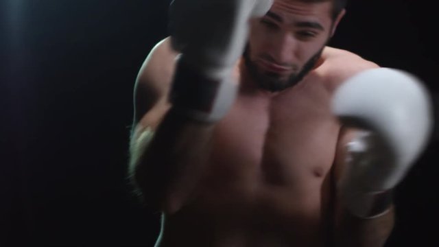 Studio shot of strong bearded boxer with bare chest throwing punches and defending head while training isolated on black background