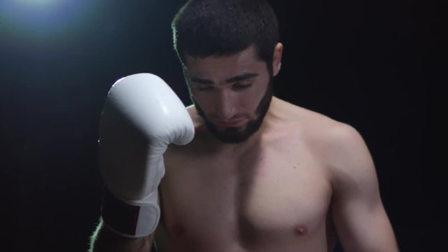 Studio shot of bearded male boxer with bare chest touching his head with boxing gloves and preparing for fight while standing isolated on black background