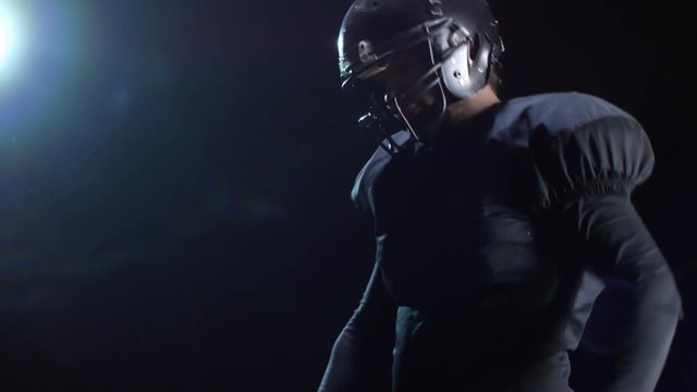 Studio shot with tilt up of American football player in helmet and full gear holding ball and warming up before practice while standing isolated on black background